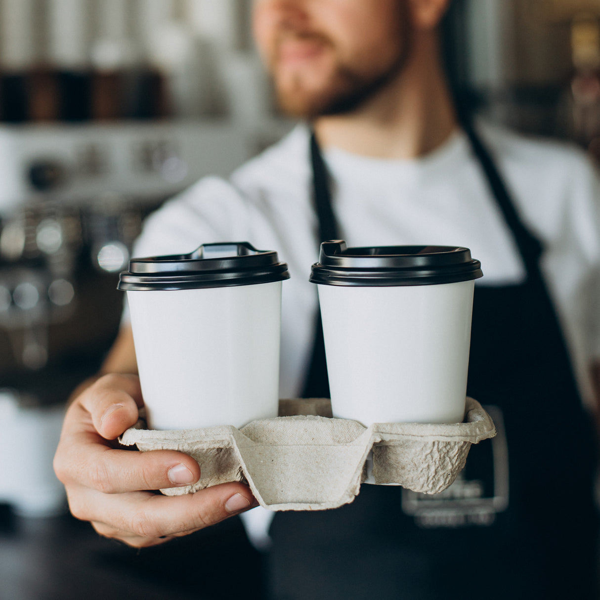 a coffee barista is handing out 2 hot cups with black lids to his customer