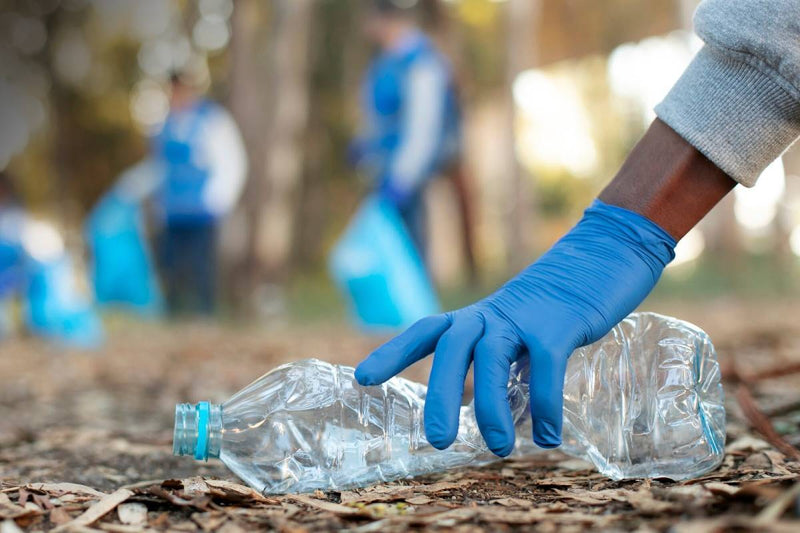 A person wearing a blue glove is picking up a crumpled plastic bottle from the. Other people are participating in a cleanup activity, holding blue trash bags.